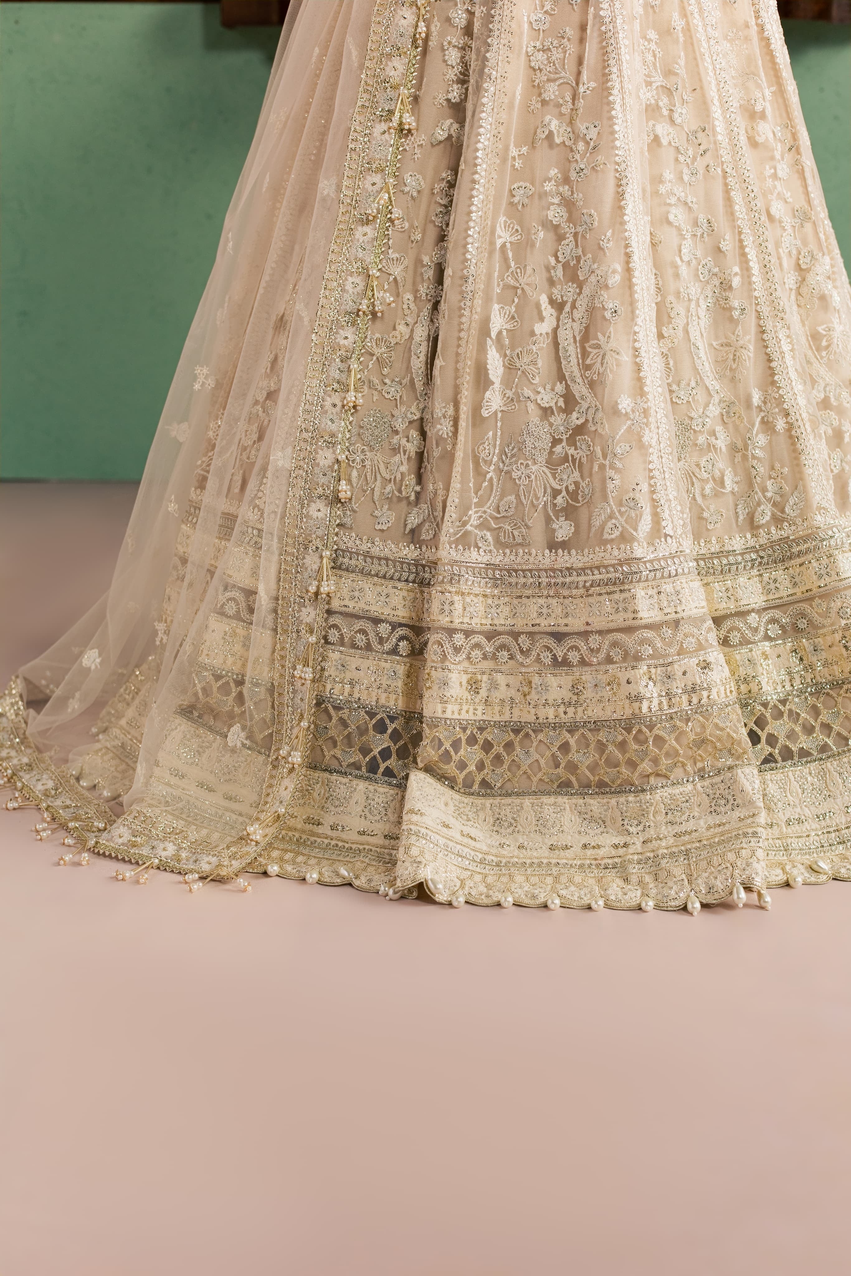 Close-up of a beige and gold heavily embroidered traditional skirt or lehenga with intricate lace, beadwork, and border detailing, shown against a soft pink floor and muted green wall.