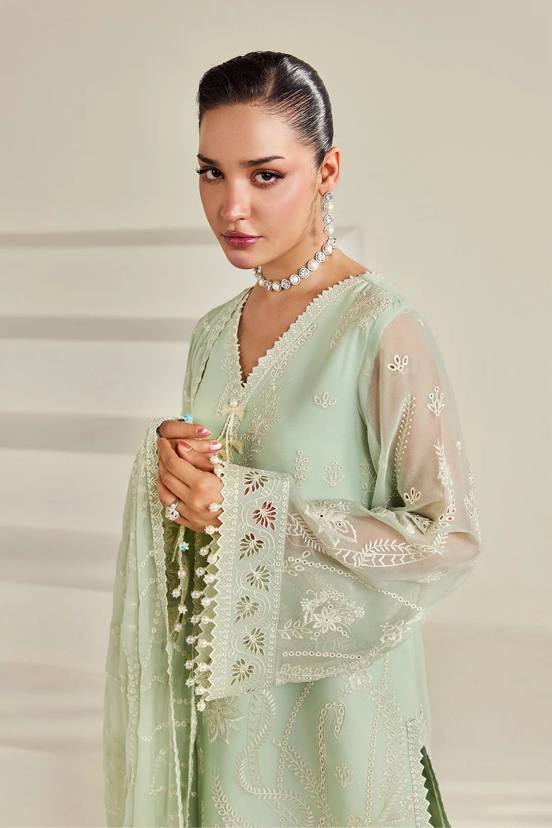 A woman modeling a light sage green traditional outfit with intricate white embroidery and sheer sleeves, styled with a matching beaded necklace and earrings against a soft beige background.