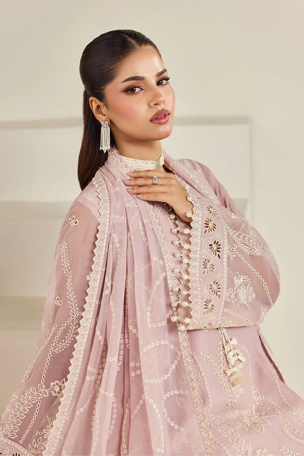 A woman models a light pink traditional outfit with intricate white embroidery and a matching sheer dupatta, set against a soft beige background.