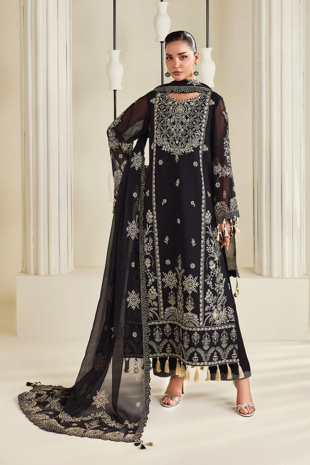 A woman models a black traditional embroidered outfit with a matching sheer dupatta, featuring intricate gold and silver embroidery, tassels, and a high neckline, set against a warm beige architectural background.