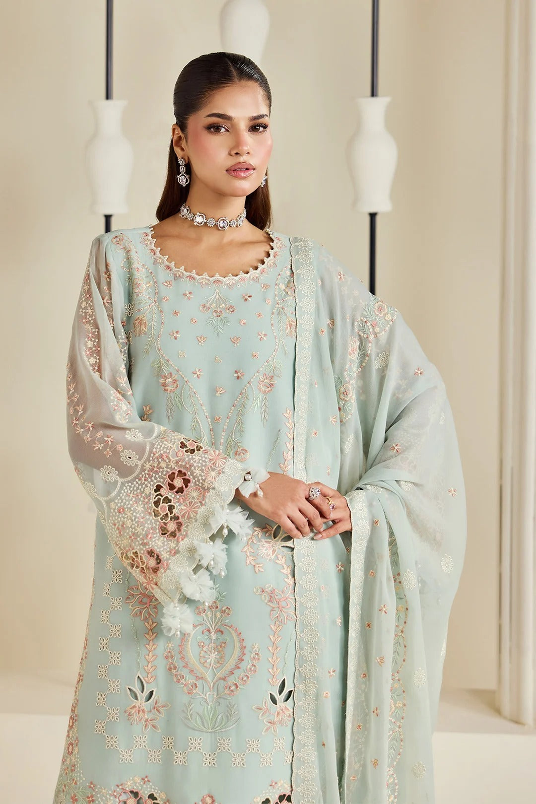 A woman models a light mint green traditional outfit with intricate floral embroidery in pink, gold, and white, featuring a matching sheer dupatta draped over her shoulders.