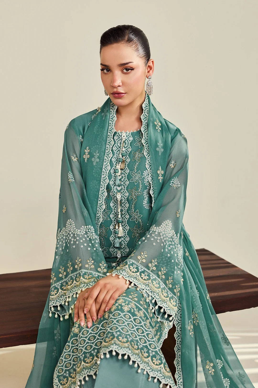 A woman models a traditional South Asian outfit consisting of a teal green embroidered kurta, matching trousers, and a sheer dupatta with intricate gold and white embroidery, seated on a wooden bench against a neutral beige background.