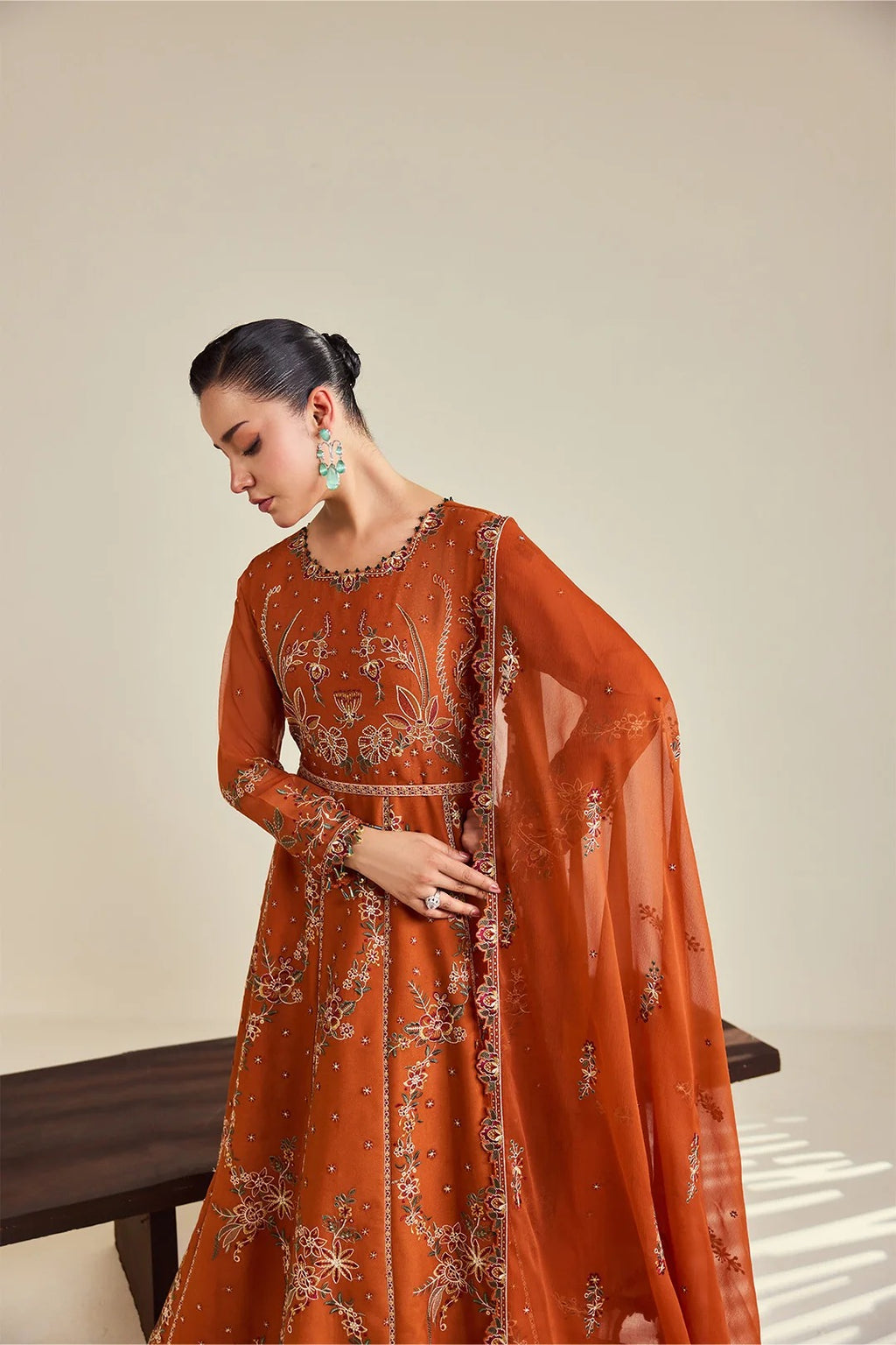 A woman models a rust-orange embroidered Anarkali gown with a matching sheer dupatta, set against a minimalist beige background.