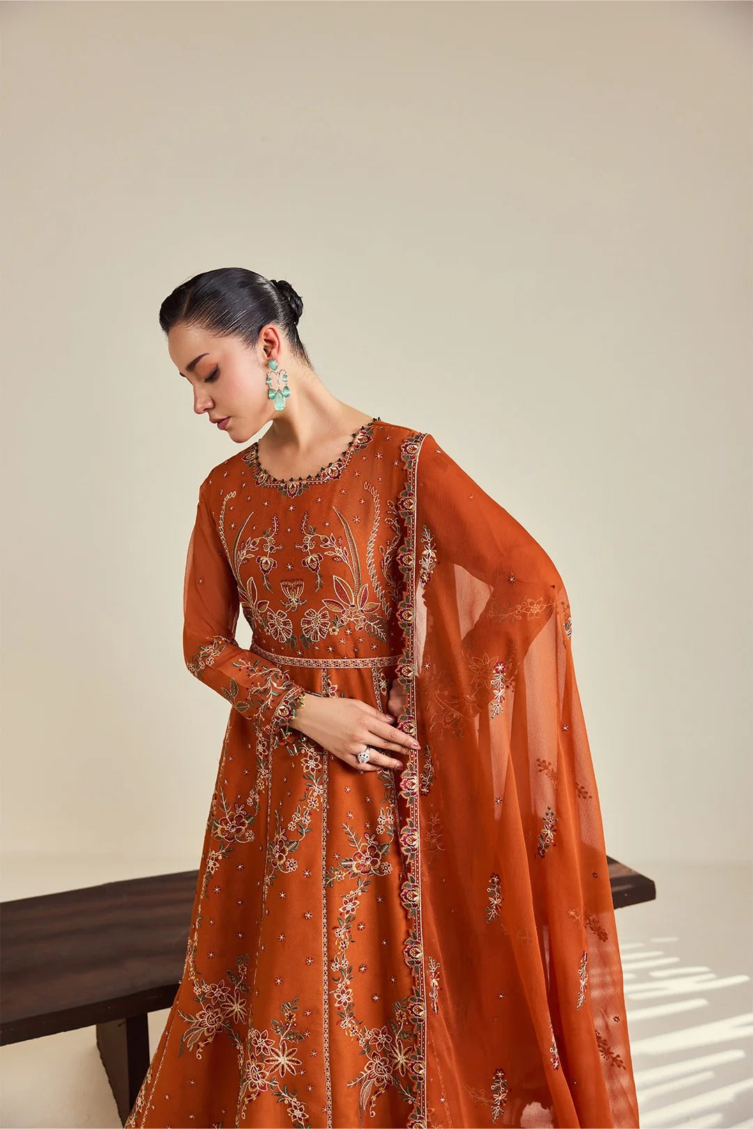 A woman models a rust-orange embroidered Anarkali gown with a matching sheer dupatta, set against a minimalist beige background.