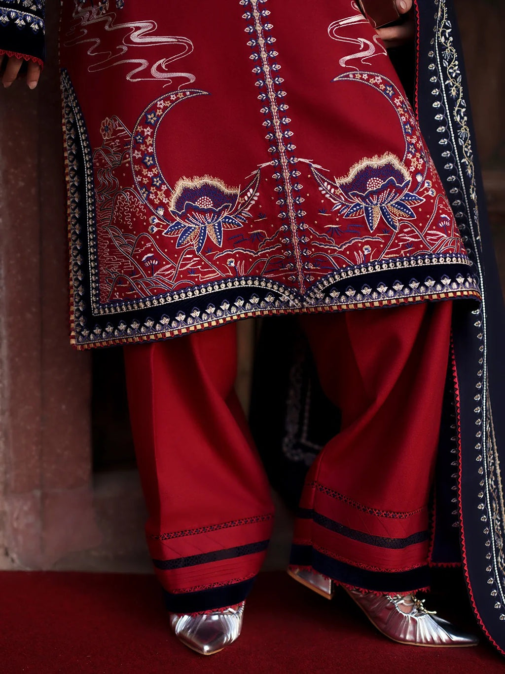 A close-up of a traditional red Indian outfit with intricate multicolor embroidery, paired with matching red pants and metallic silver shoes.