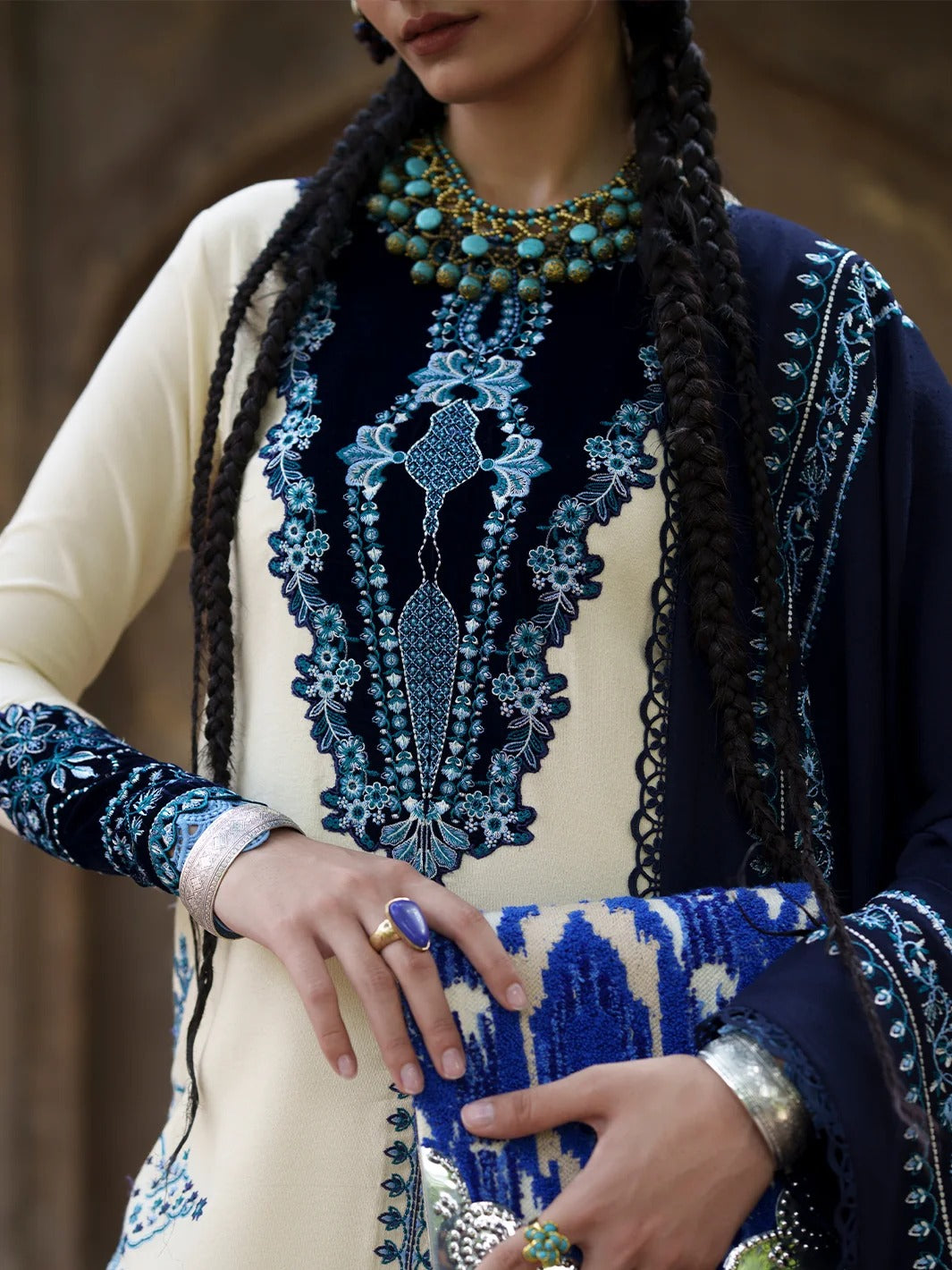 A close-up of a model wearing a traditional South Asian outfit with a cream-colored kurta featuring intricate blue and navy embroidery, a navy blue dupatta with matching embroidery, and a blue patterned clutch bag.