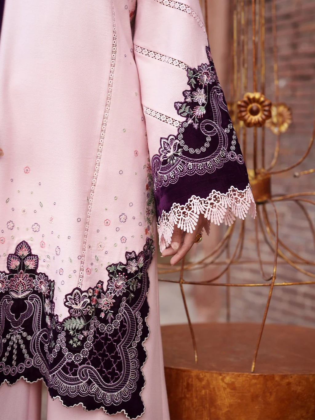 A close-up of a light pink traditional outfit with dark purple embroidered lace and floral detailing on the sleeve and hem, shown on a mannequin or model in an indoor styled setting.