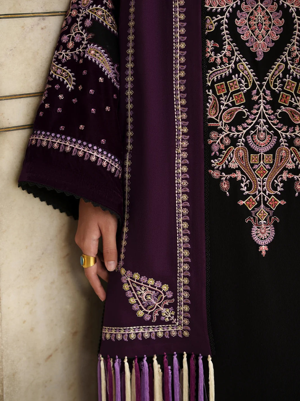 A close-up of a traditional embroidered garment or textile in deep purple and black, featuring ornate floral and paisley embroidery in pink, gold, and white, with a hand holding the purple sleeve.