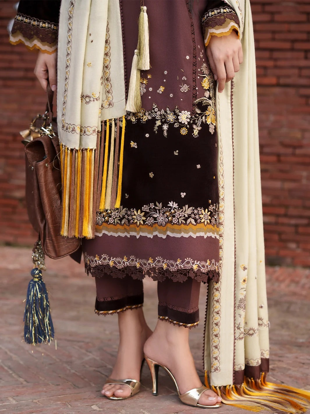 A person wearing traditional South Asian attire, featuring a dark brown embroidered kurta with gold and white floral patterns, matching brown trousers, a cream dupatta with gold tassels, and metallic gold high-heeled sandals, set against a rustic brick wall background.