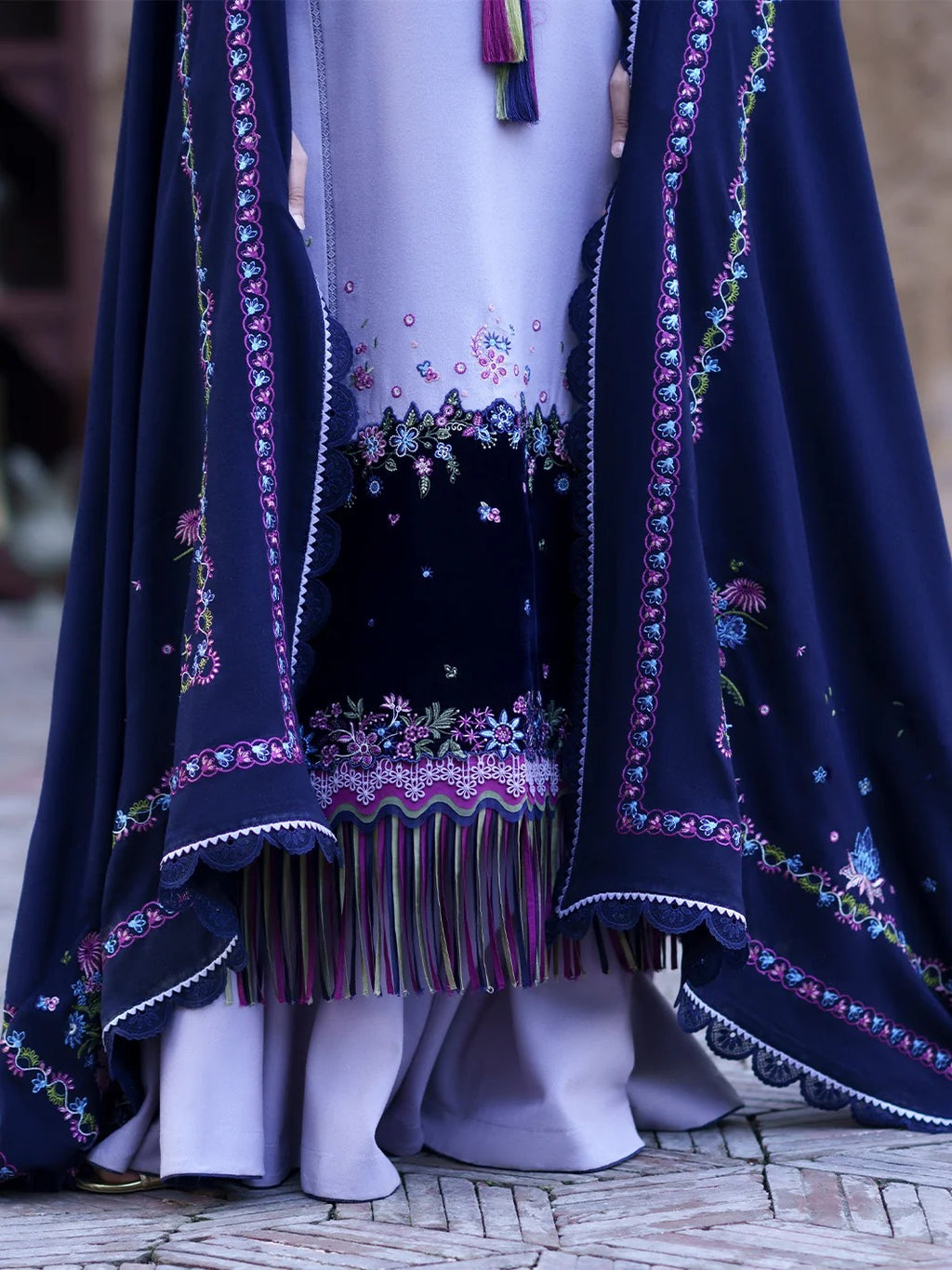 A close-up of a traditional embroidered outfit with a navy blue outer layer draped over a light lavender dress. The fabric features intricate floral embroidery in pink, purple, and green, with a striped fringe detail at the hem.
