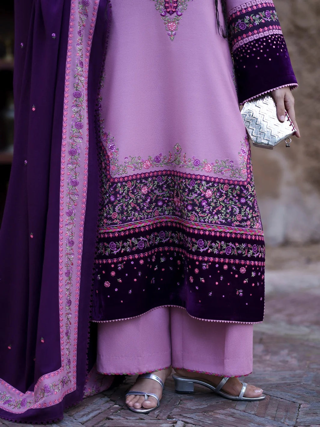 A close-up of a person wearing a lavender and deep purple embroidered traditional outfit with a matching dupatta, holding a small silver clutch.