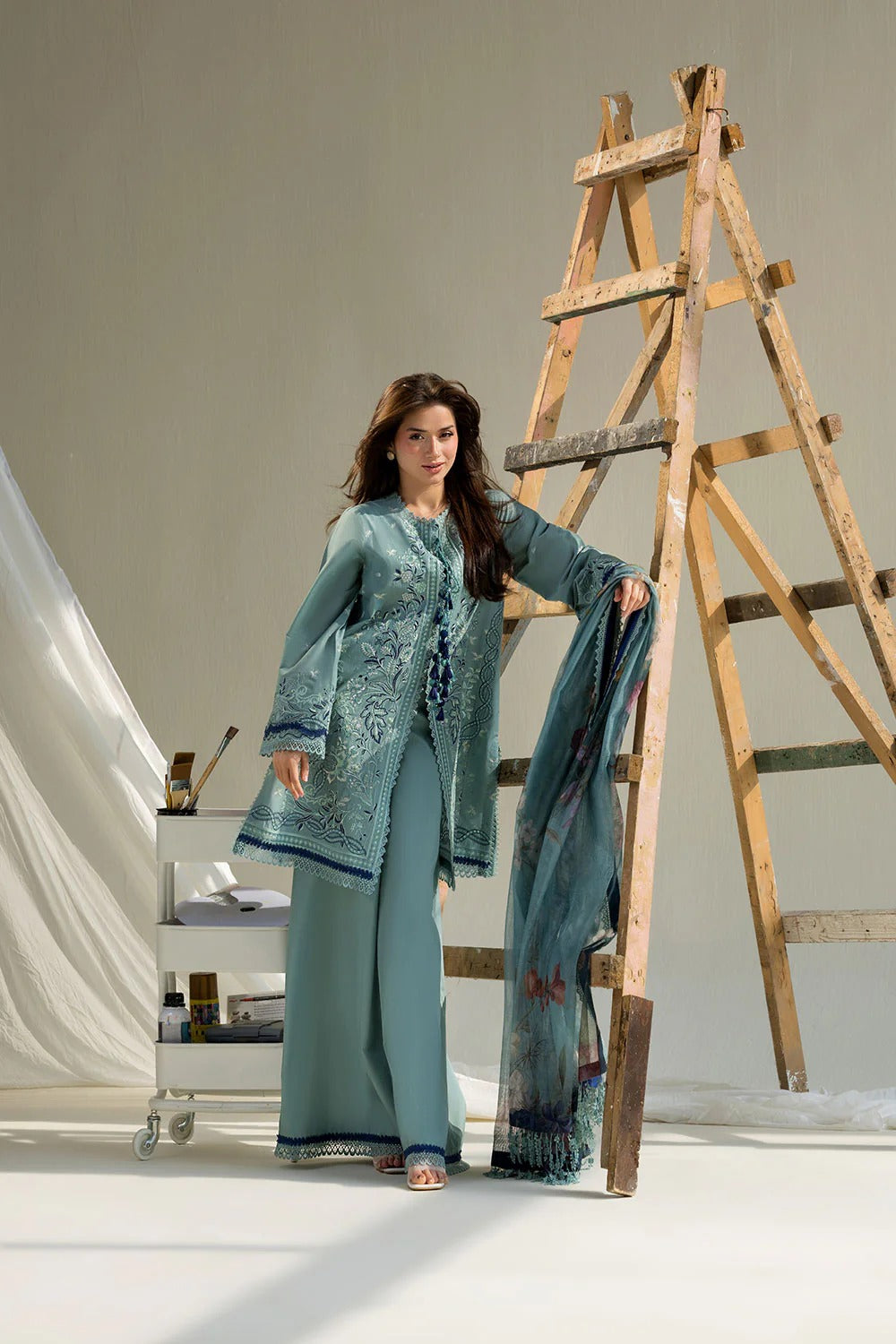 Woman in a teal outfit standing next to a wooden ladder in a studio setting.