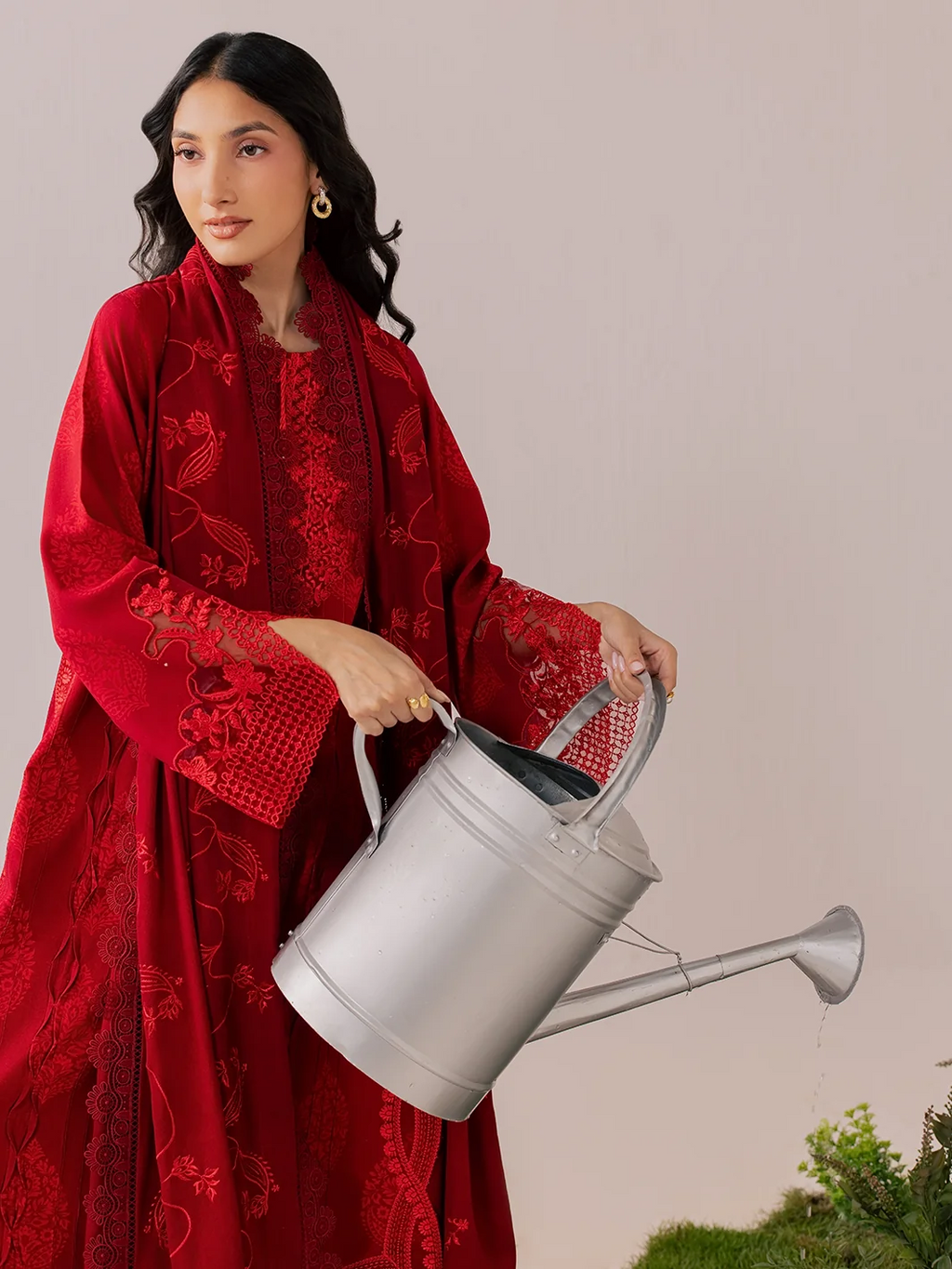 A woman wearing a long-sleeved red embroidered dress holds a silver metal watering can against a plain beige background.
