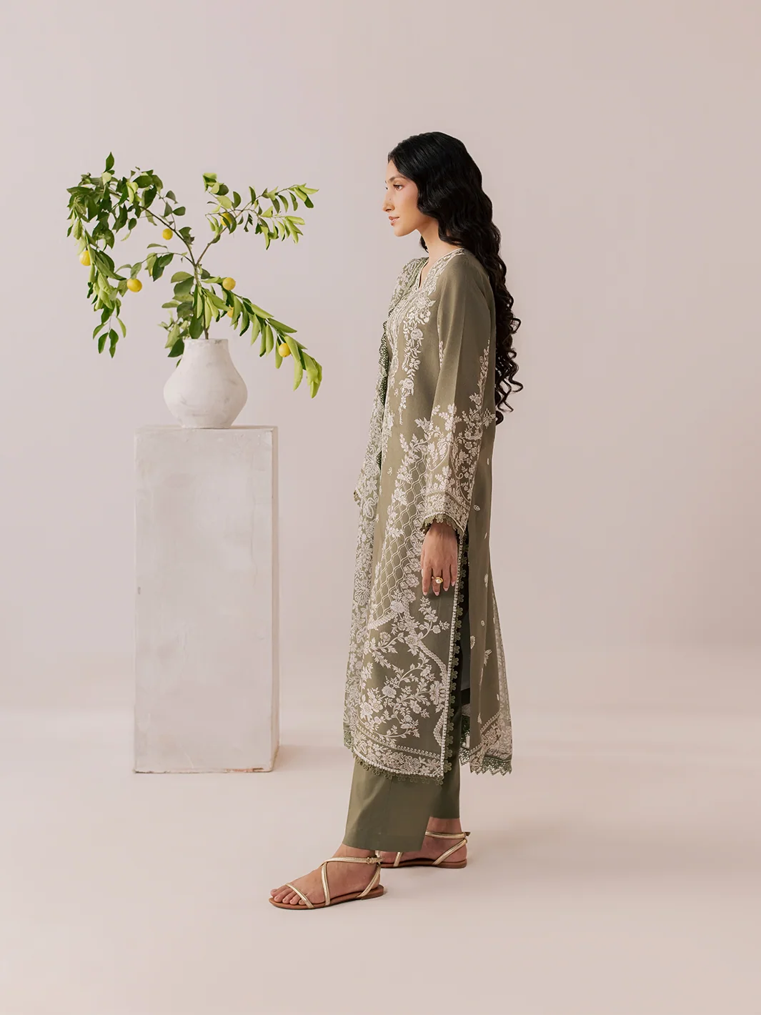 A woman stands in profile wearing an olive green embroidered kurta with matching wide-leg pants and strappy flat sandals, posed beside a white pedestal holding a vase with green branches against a soft beige studio background.