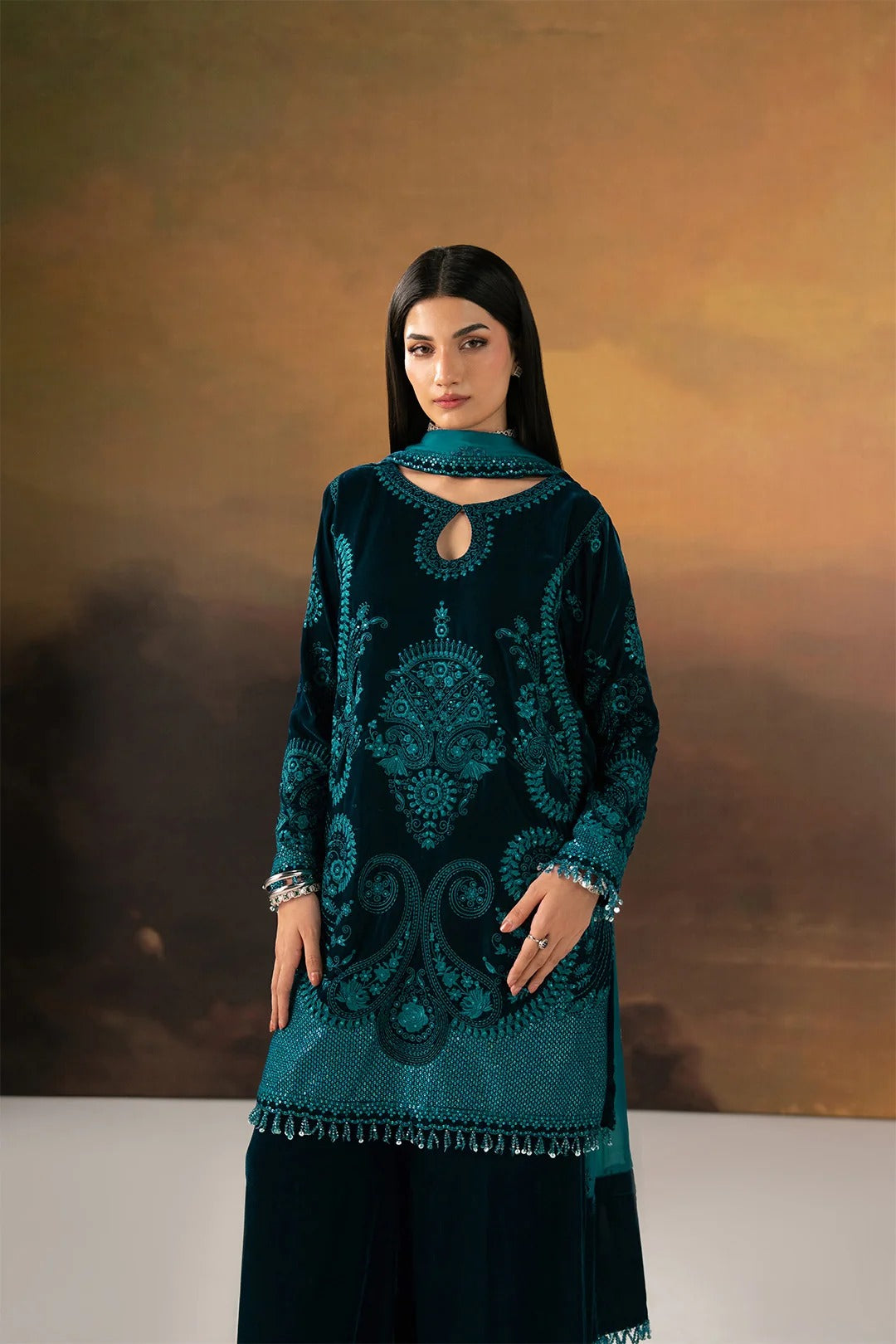 A woman modeling a dark teal embroidered kurta with a matching dupatta and black wide-leg pants, set against a warm brown studio backdrop.