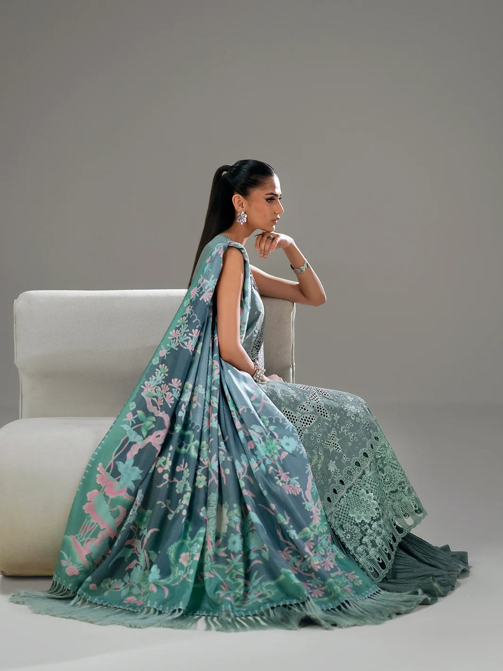 Woman in a green floral saree sitting on a white chair against a gray background