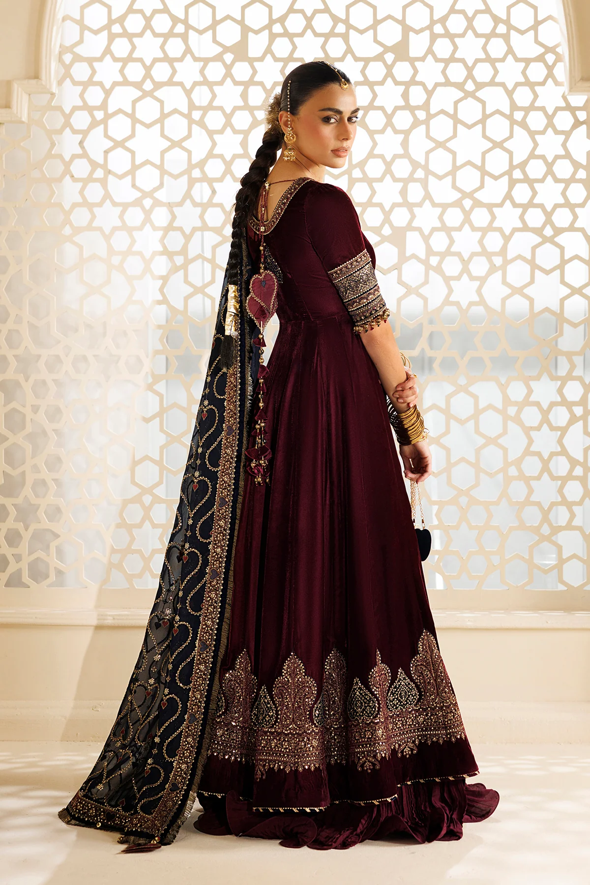 A woman modeling a deep maroon Anarkali gown with ornate gold embroidery and a black dupatta featuring gold patterns, posed in front of a white geometric screen.
