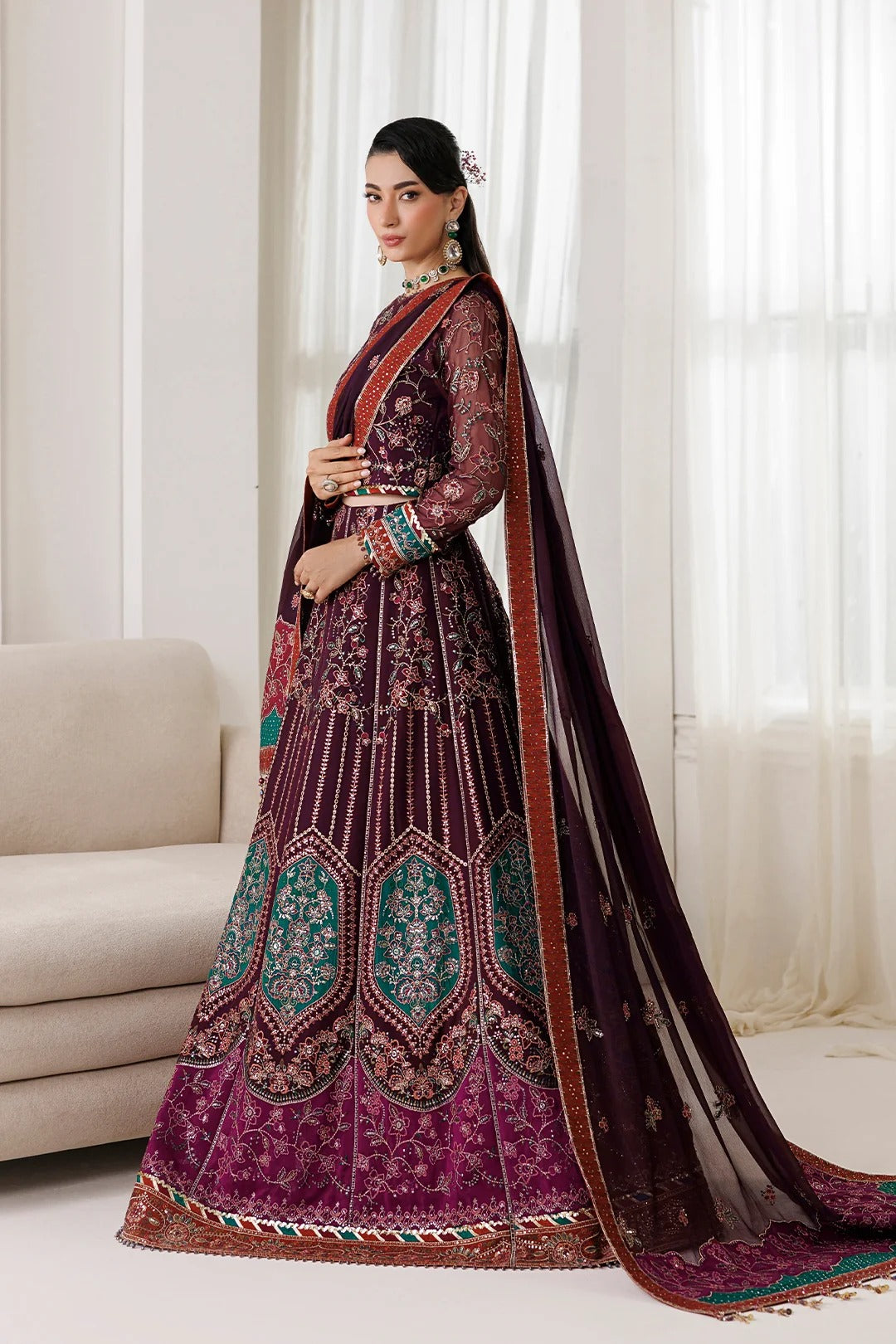 A woman models a traditional Indian anarkali gown in deep purple with intricate gold embroidery and teal accents, paired with a matching sheer dupatta, in an indoor setting with a sofa and white curtains.