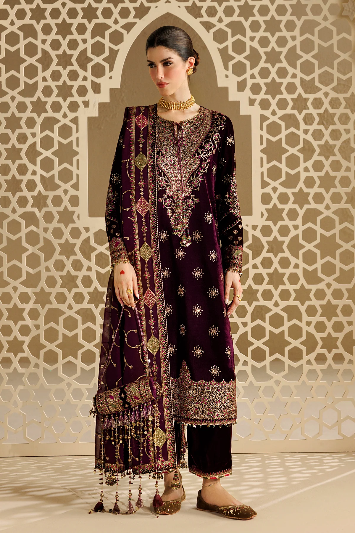 A woman models a traditional South Asian outfit consisting of a deep purple velvet kurta with intricate gold and silver embroidery, matching trousers, and a coordinating dupatta, set against a beige geometric patterned wall.