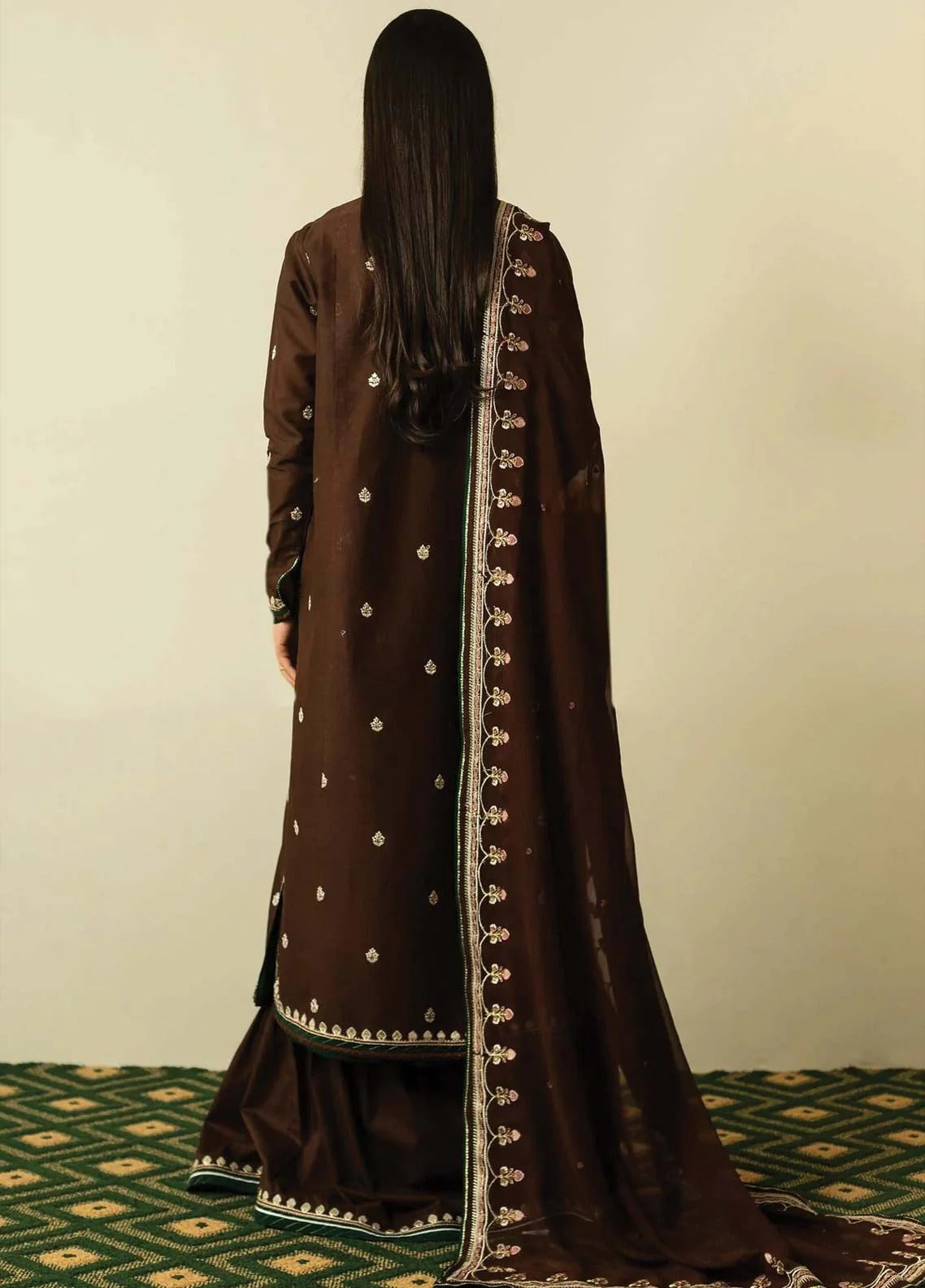 A woman viewed from behind wearing a dark brown traditional outfit with silver embroidered details and a matching dupatta, standing against a plain beige wall on a patterned green carpet.