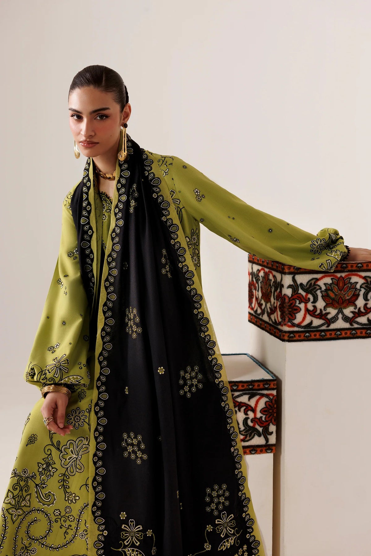 A close-up of a woman wearing a traditional embroidered kurta in olive green with black floral embroidery, paired with a black dupatta featuring gold and black border detailing, accessorized with gold jewelry.