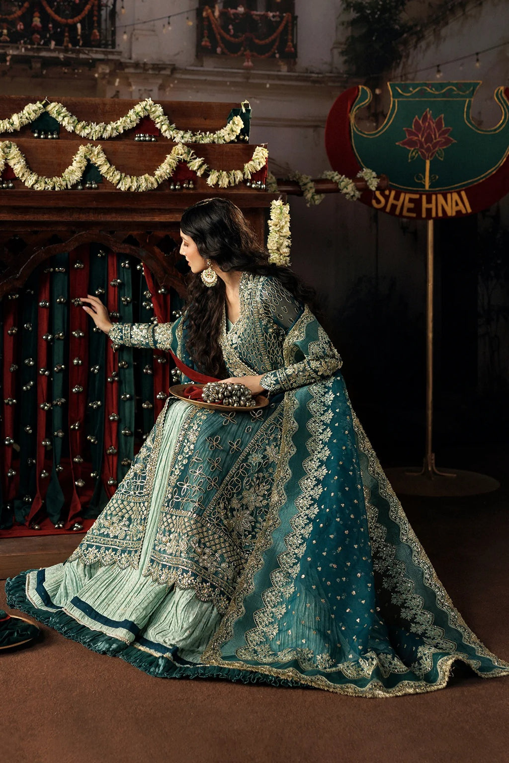 A woman wearing an ornate teal and silver embroidered traditional outfit poses indoors in front of decorated curtains and garlands. A sign reading SHEHNAI is visible in the background.