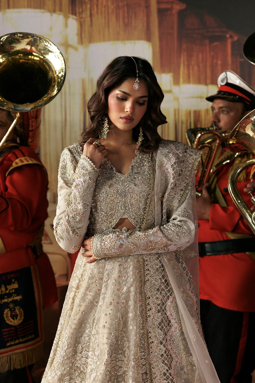 A woman wearing an ornate silver and white embroidered traditional outfit with a sheer draped overlay, standing in front of a warm-toned backdrop with band members holding brass instruments visible on both sides.