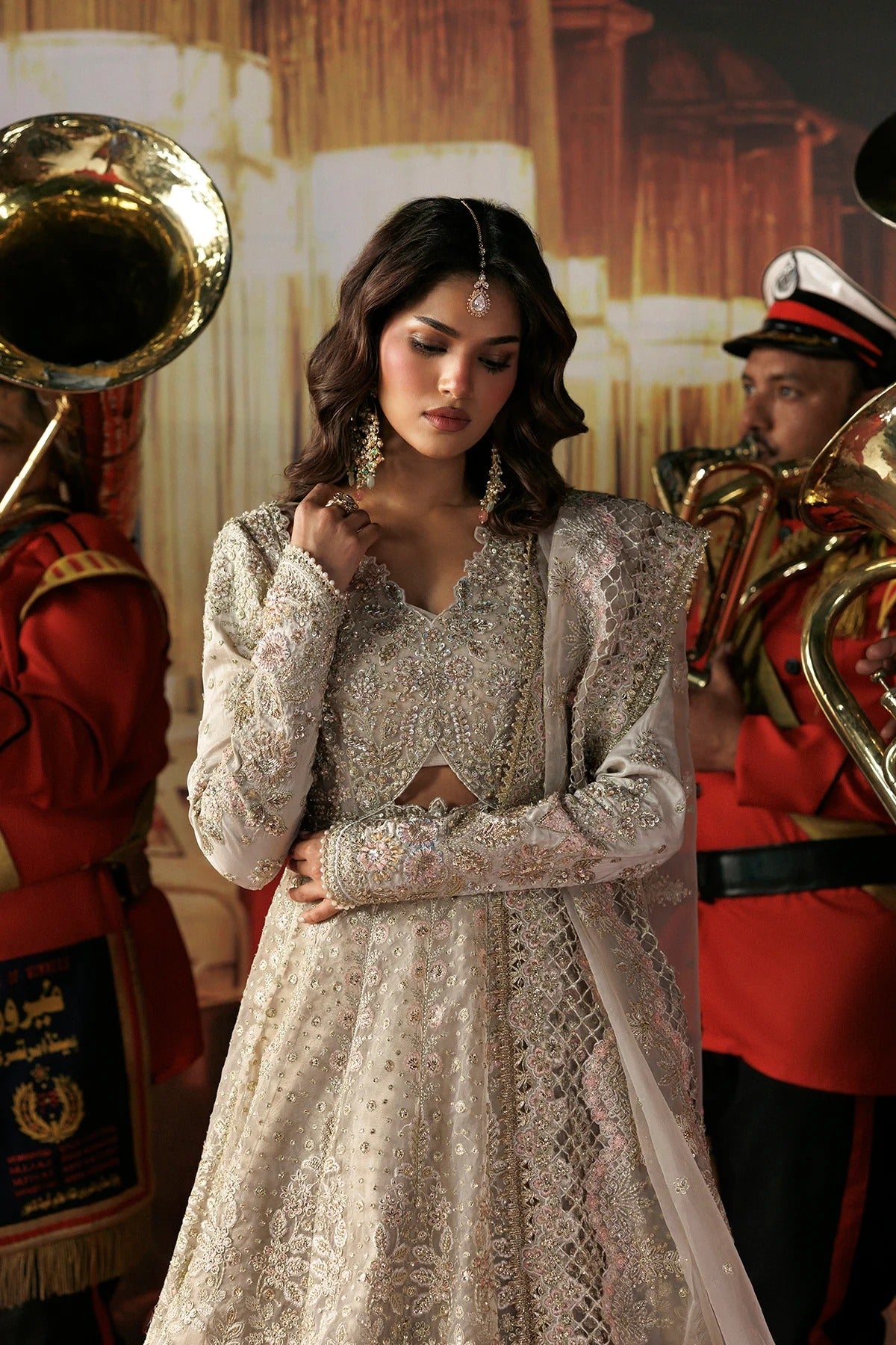 A woman wearing an ornate silver and white embroidered traditional outfit with a sheer draped overlay, standing in front of a warm-toned backdrop with band members holding brass instruments visible on both sides.
