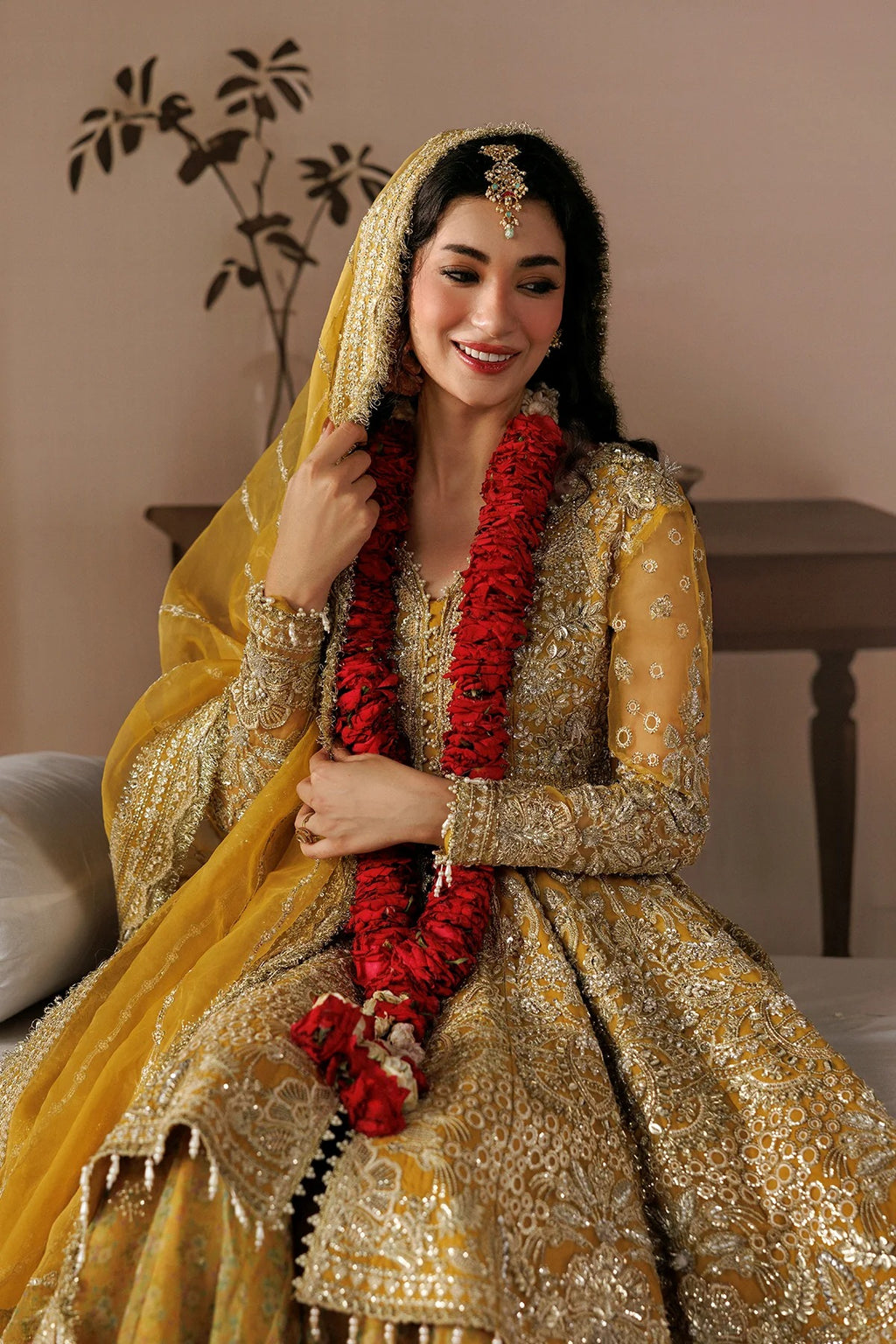 A woman wearing an ornate gold and yellow traditional bridal outfit with intricate embroidery, a matching veil, and a red flower garland, seated indoors against a warm beige wall.