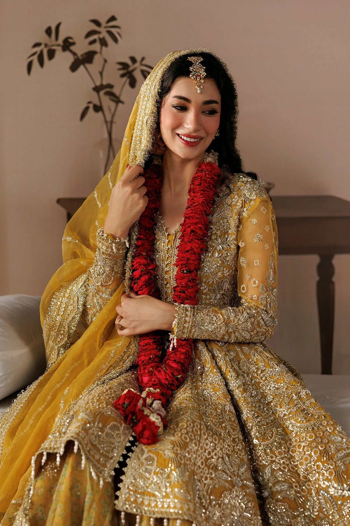 A woman wearing an ornate gold and yellow traditional bridal outfit with intricate embroidery, a matching veil, and a red flower garland, seated indoors against a warm beige wall.