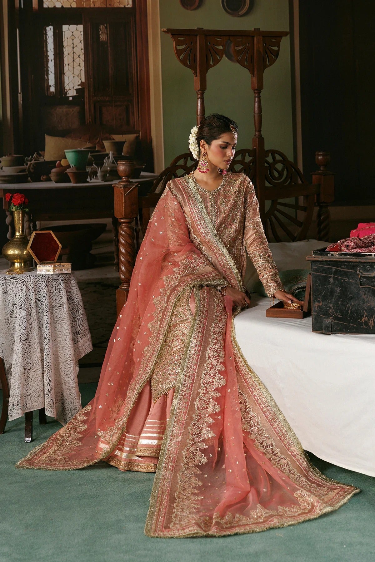 A woman wearing an ornate peach-pink and gold embroidered traditional outfit with a sheer dupatta, posed in a vintage-style room with wooden furniture and a bed.