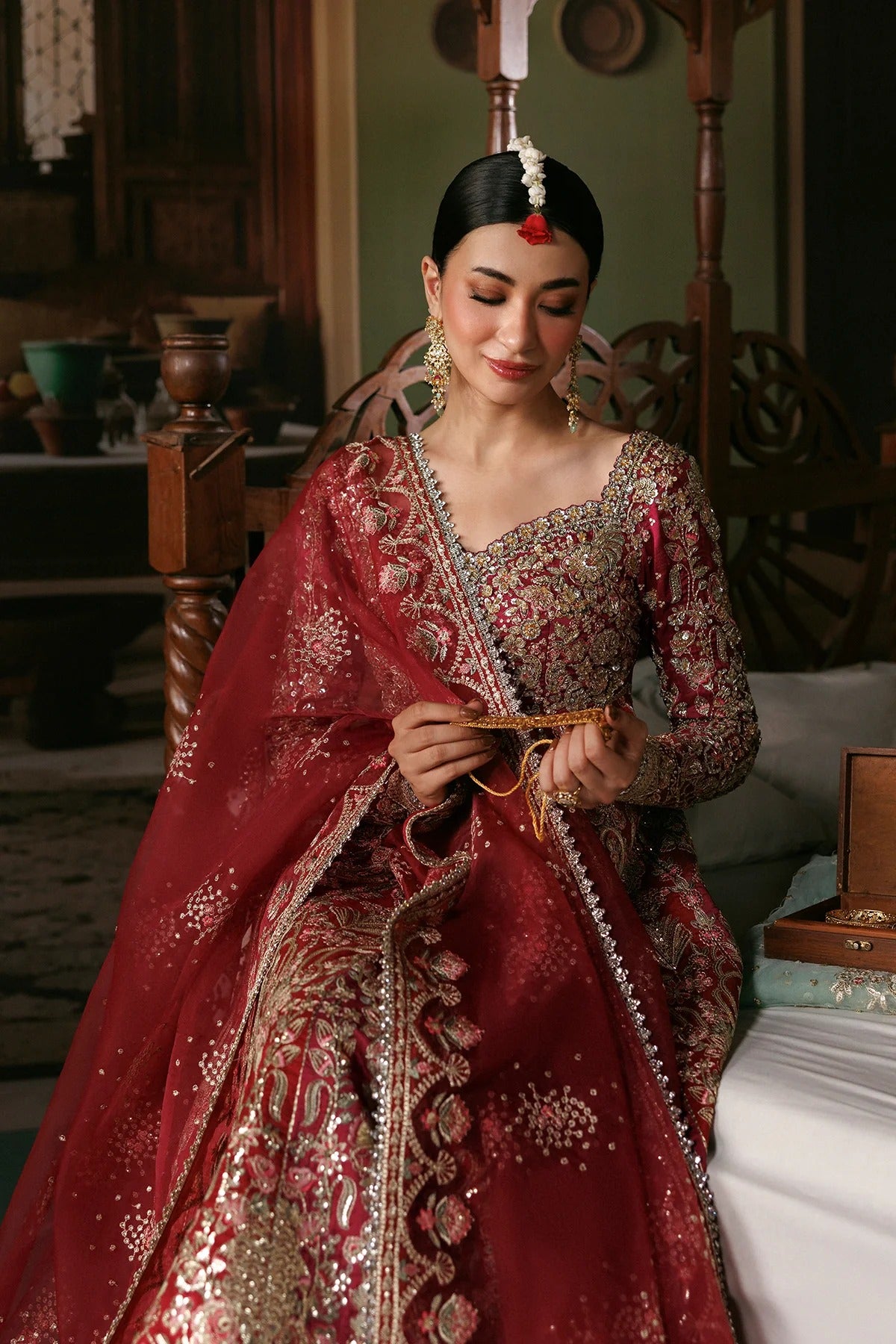 A woman wearing an ornate red and gold embroidered traditional bridal outfit with a sheer dupatta, seated indoors in a decorated room.