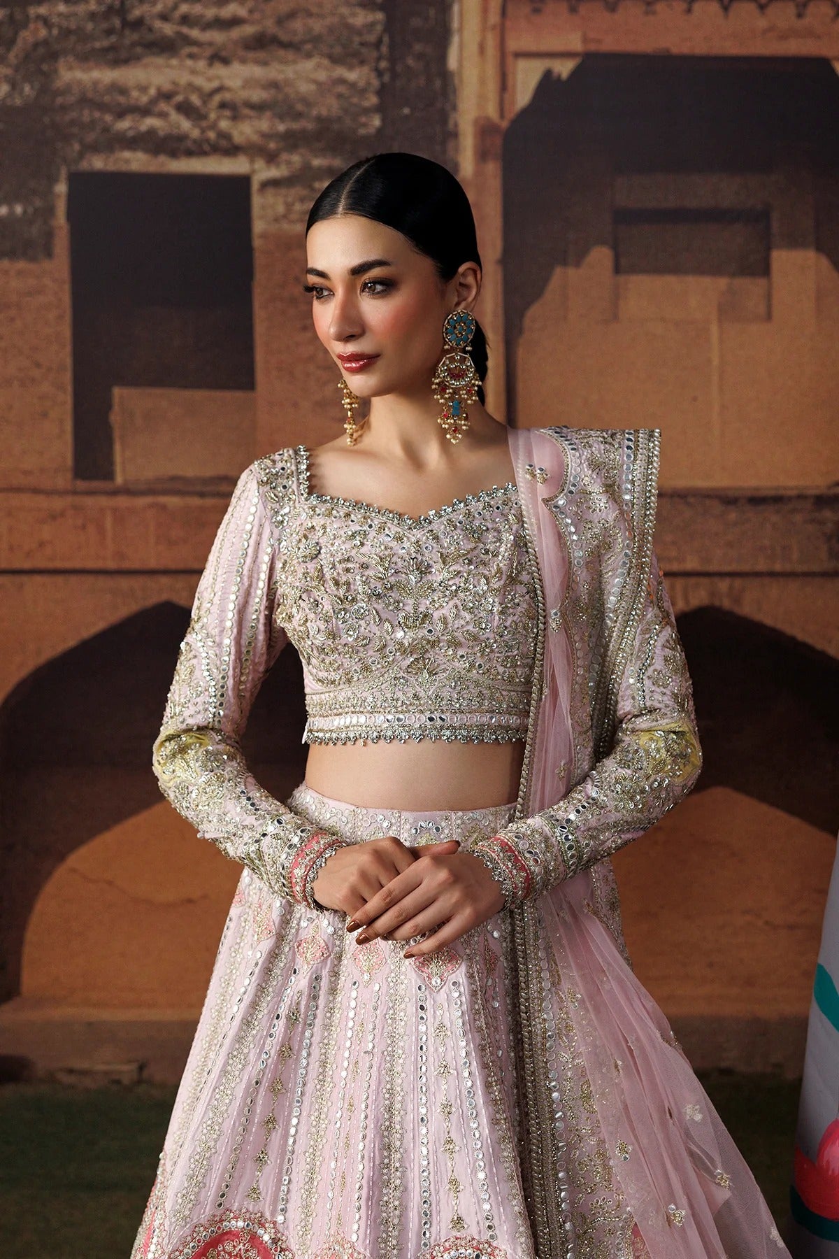 A woman wearing an ornate light pink and silver embellished traditional outfit with a matching dupatta, standing in front of a warm-toned architectural backdrop.