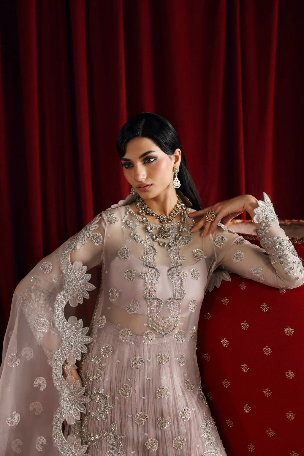 A woman wearing an ornate light pink traditional embroidered gown with silver embellishments, posed against a deep red velvet curtain and matching red upholstered furniture.