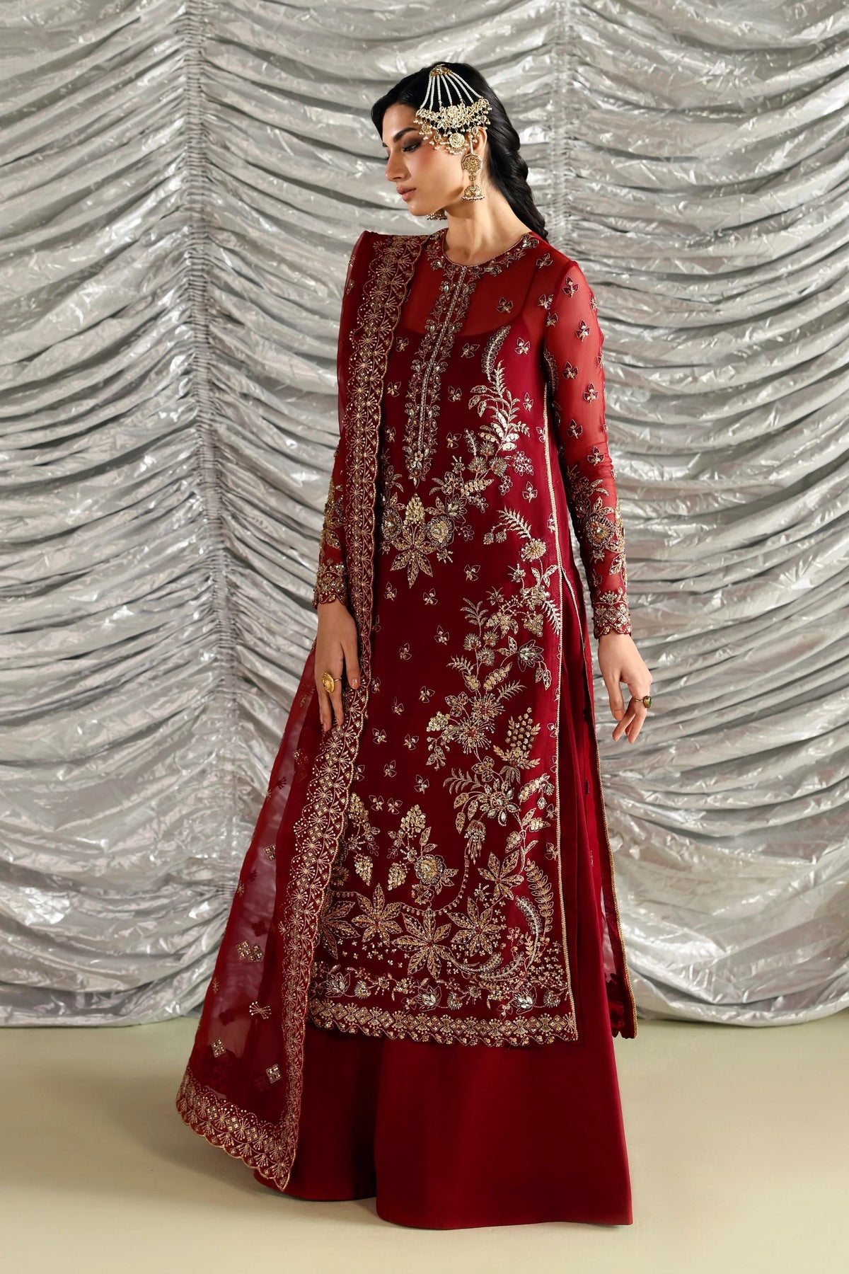 A woman models a traditional deep red Indian salwar kameez suit with intricate gold embroidery, a matching dupatta, and a silver draped backdrop.