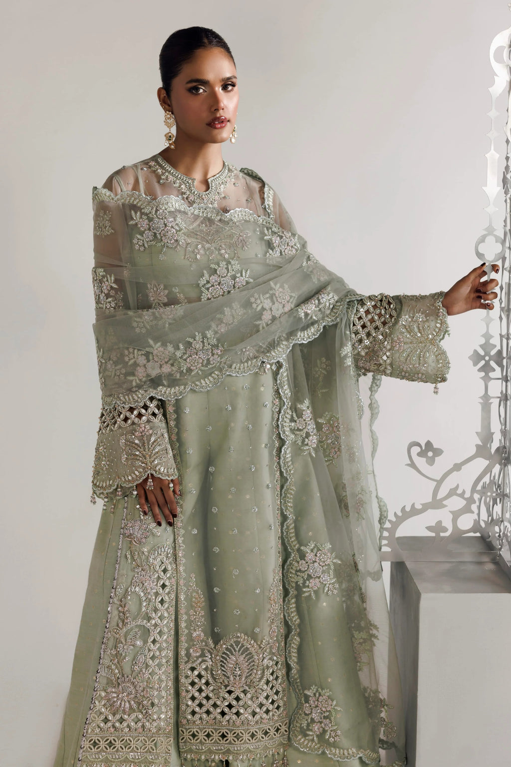A woman models a traditional Indian outfit consisting of a sage green anarkali kurta with intricate silver embroidery, a matching dupatta, and a sheer long-sleeve blouse. She is standing in a studio setting with a light gray background and a decorative metal screen on the right.