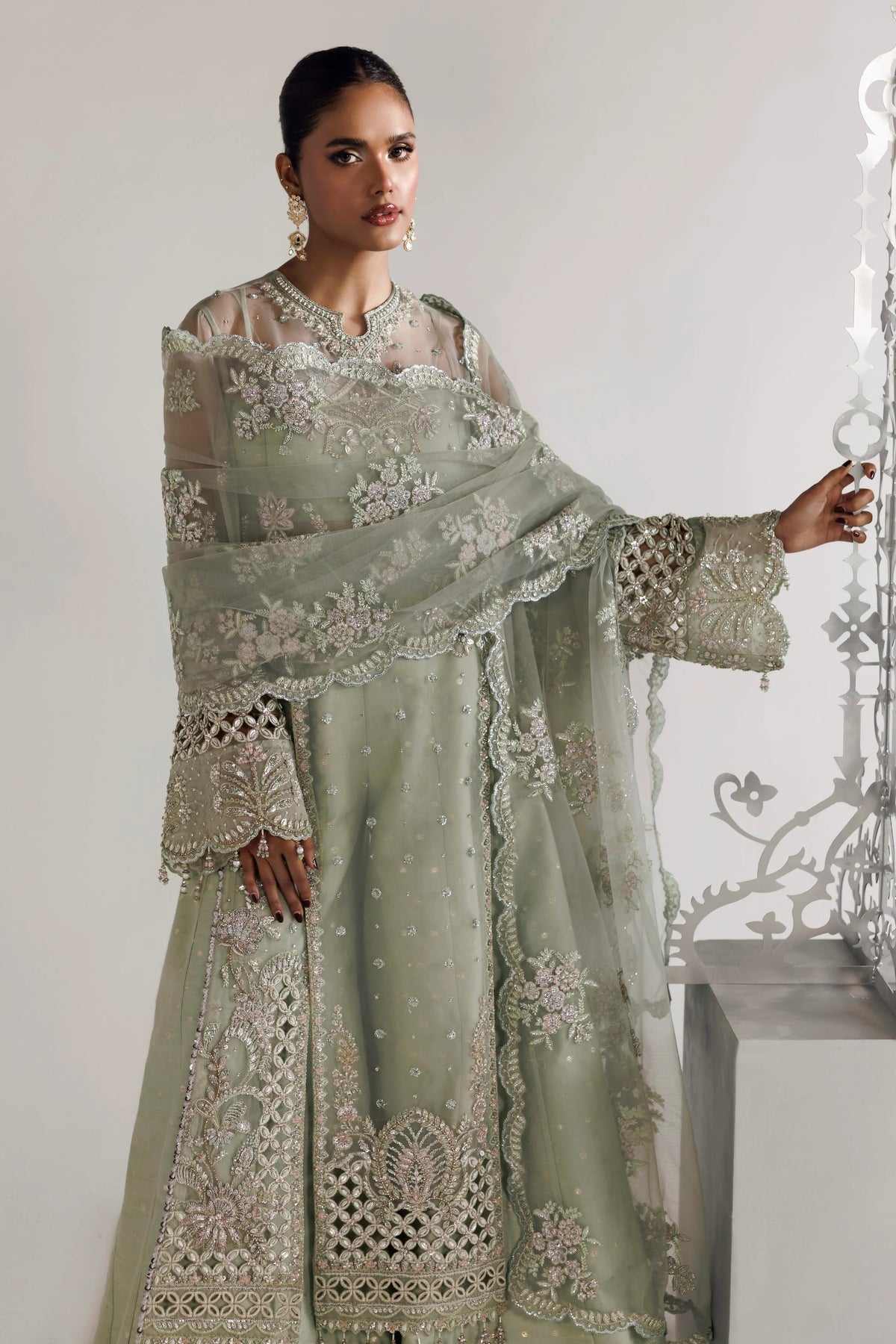 A woman models a traditional Indian outfit consisting of a sage green anarkali kurta with intricate silver embroidery, a matching dupatta, and a sheer long-sleeve blouse. She is standing in a studio setting with a light gray background and a decorative metal screen on the right.