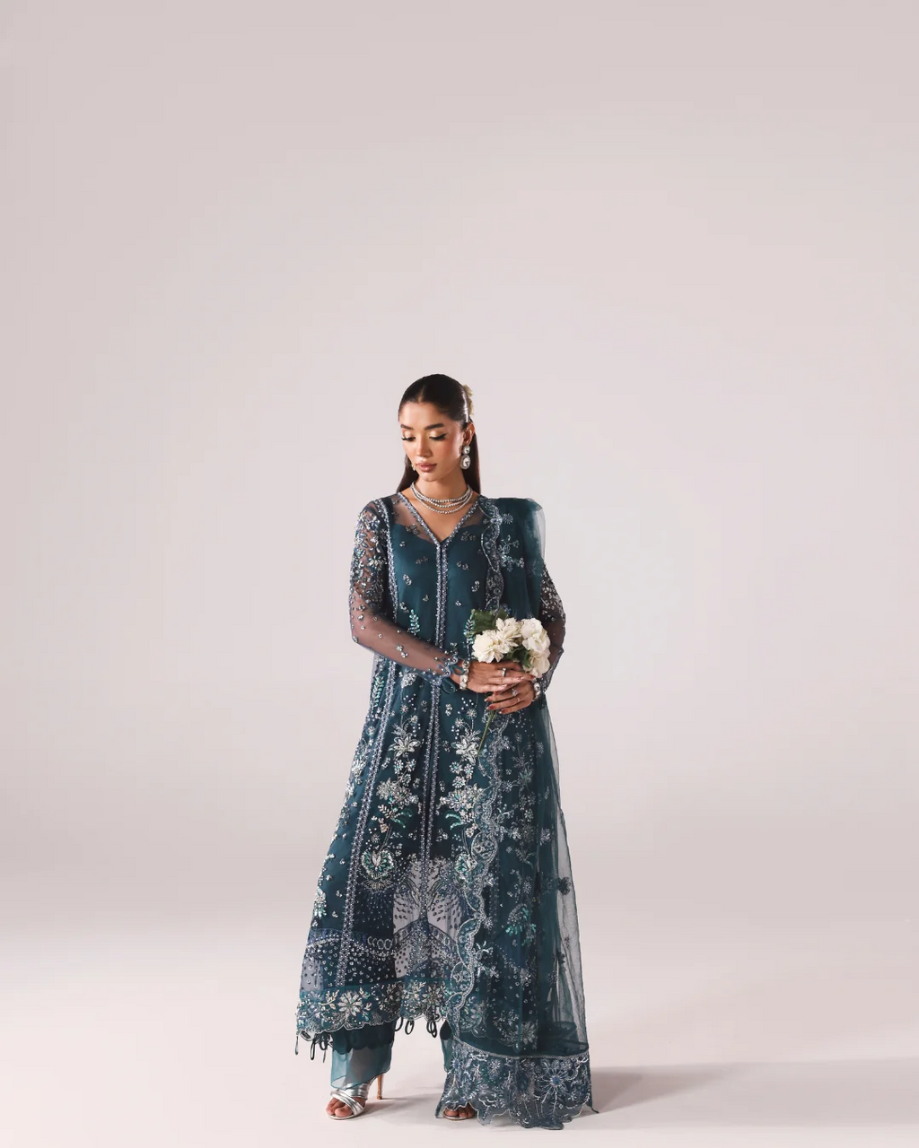 A woman modeling a dark teal blue embroidered traditional outfit with sheer sleeves and a matching dupatta, holding a small bouquet of white flowers against a plain light gray studio background.