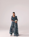 A woman modeling a dark teal blue embroidered traditional outfit with sheer sleeves and a matching dupatta, holding a small bouquet of white flowers against a plain light gray studio background.