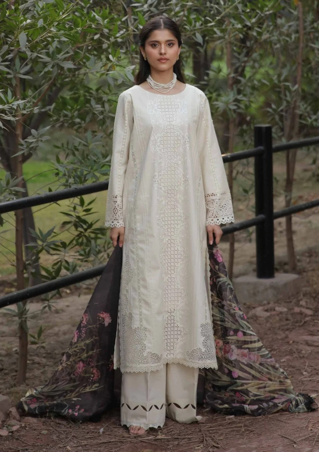 A woman stands outdoors in a wooded area wearing a cream-colored traditional outfit with a long embroidered kurta, matching pants, and a dark patterned dupatta.