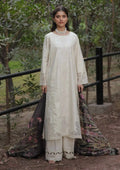 A woman stands outdoors in a wooded area wearing a cream-colored traditional outfit with a long embroidered kurta, matching pants, and a dark patterned dupatta.