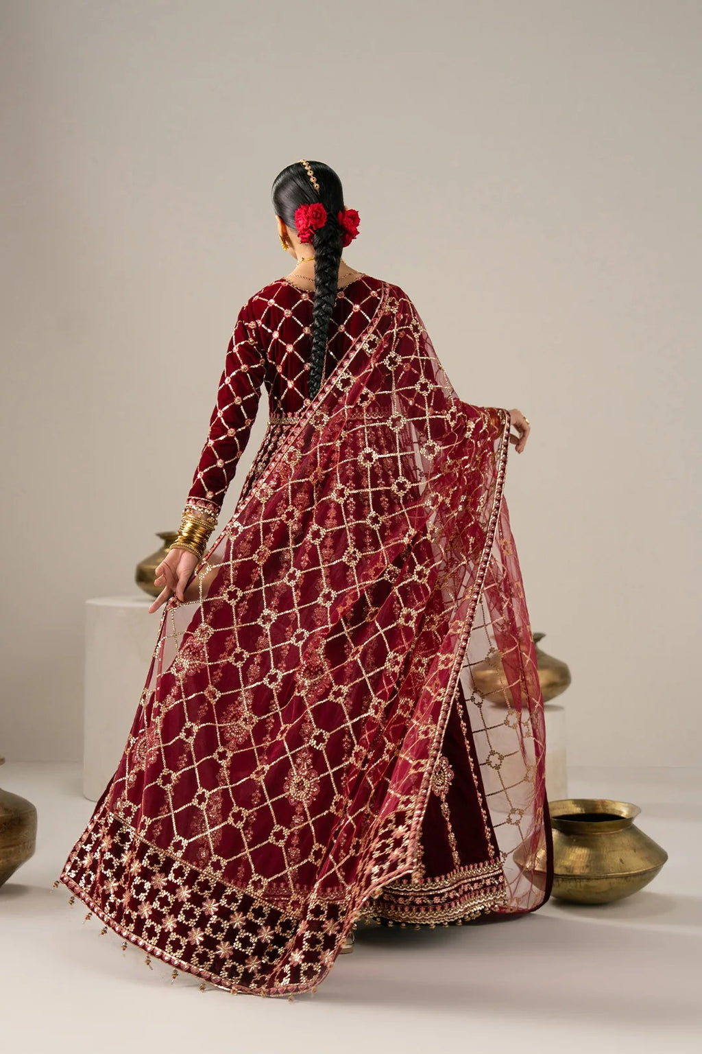 A woman shown from behind wearing a deep maroon red saree with intricate gold geometric embroidery and a matching embroidered blouse. She is standing in a studio setting with neutral beige walls and decorative brass pots on the floor.