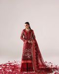 A woman models a traditional red embroidered Indian outfit with a matching dupatta, standing on a white studio floor scattered with red rose petals.