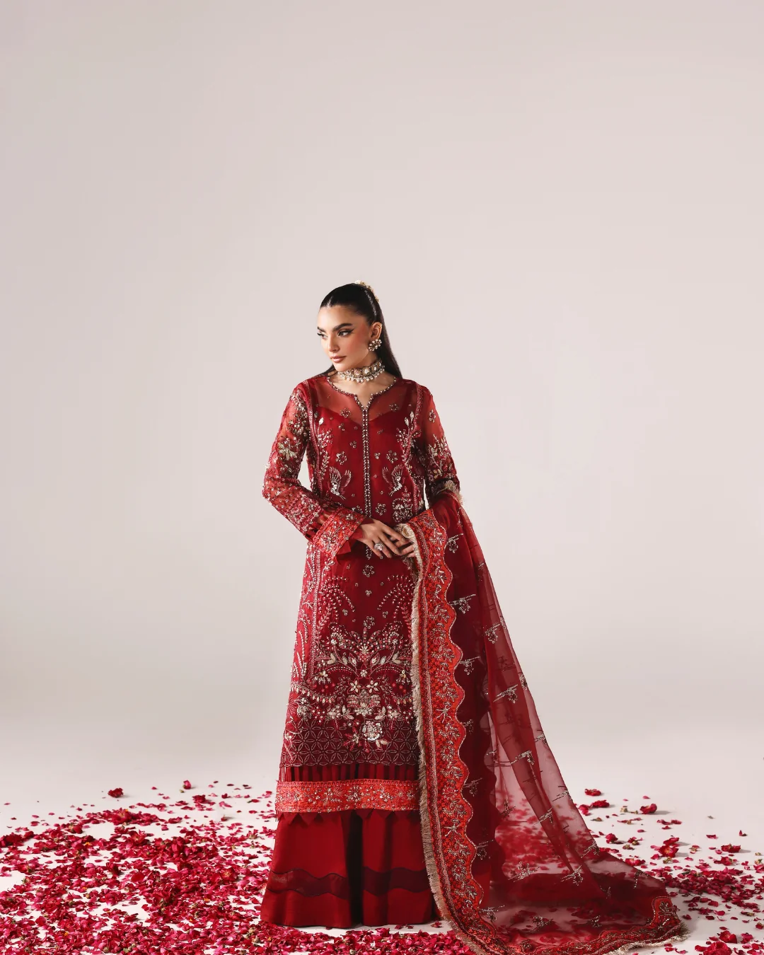 A woman models a traditional red embroidered Indian outfit with a matching dupatta, standing on a white studio floor scattered with red rose petals.