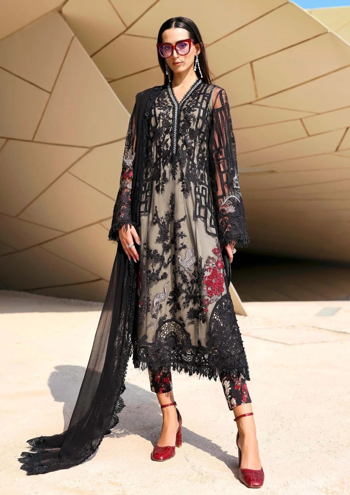 Woman wearing a long, dark floral dress with sheer overlay in front of a modern architectural background.