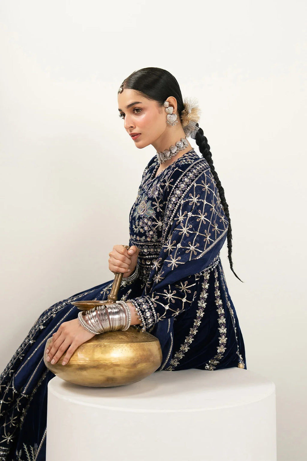 A woman in a navy blue embroidered traditional outfit sits on a white pedestal, holding a brass singing bowl against a plain light background.