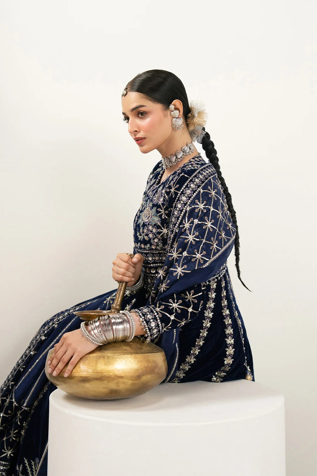 A woman in a navy blue embroidered traditional outfit sits on a white pedestal, holding a brass singing bowl against a plain light background.