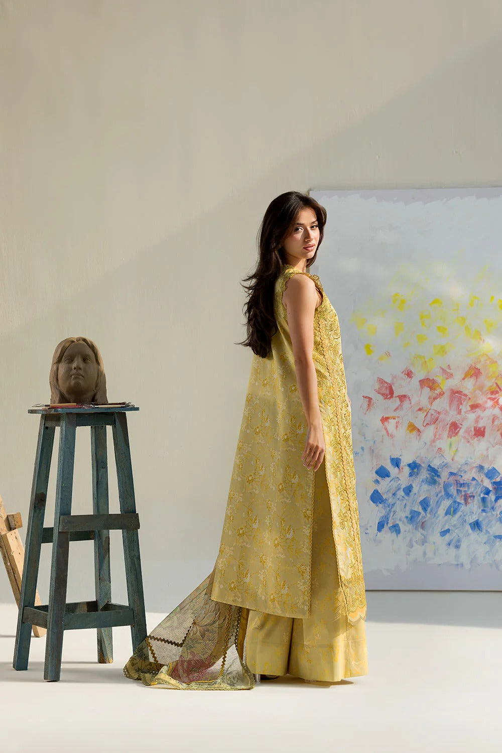 Woman in a yellow traditional outfit standing next to a stool with a small sculpture on it, against a neutral background.