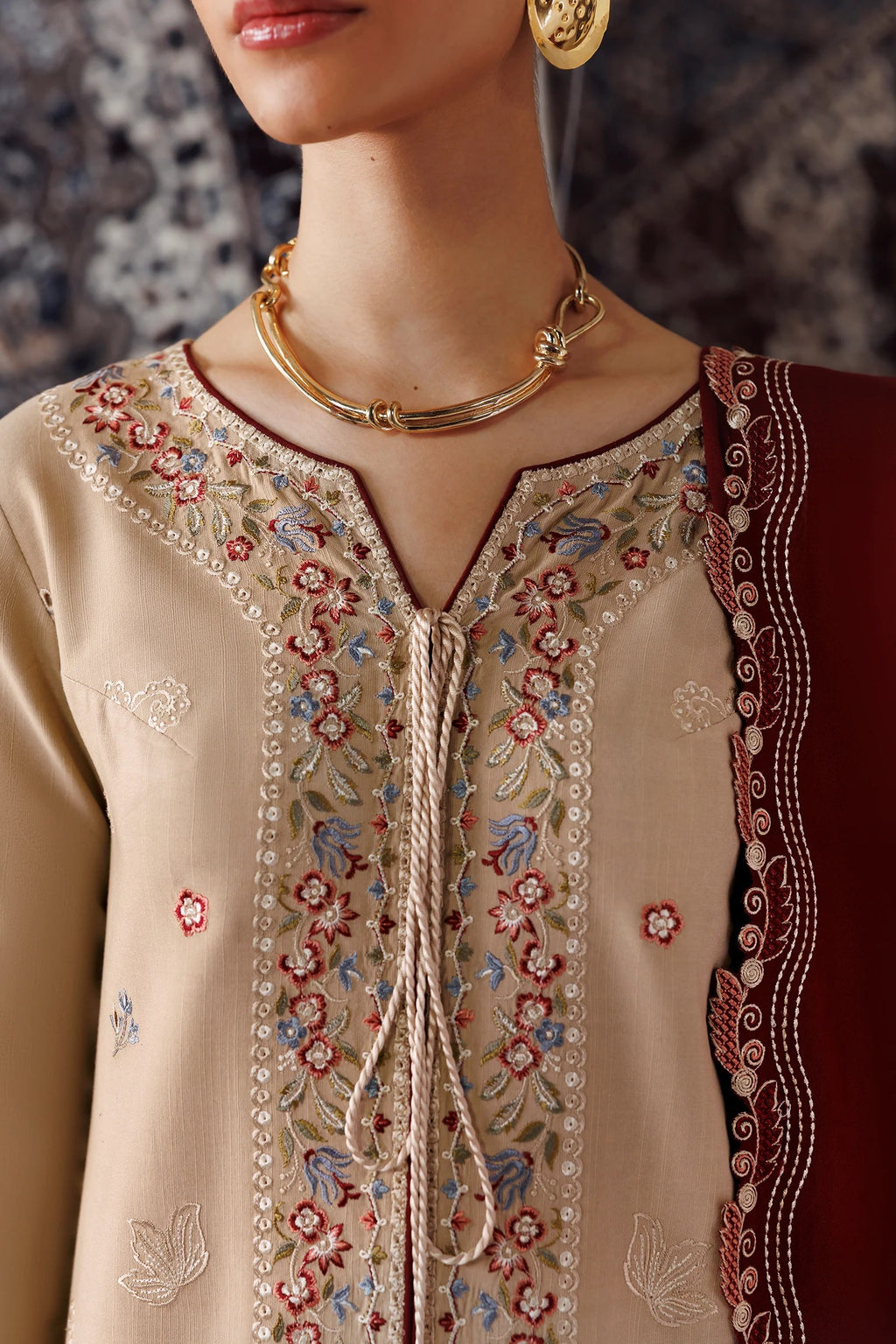 Close-up of a beige embroidered kurta with floral embroidery, a drawstring neckline, and a deep red dupatta draped over one shoulder, worn by a model with gold jewelry.