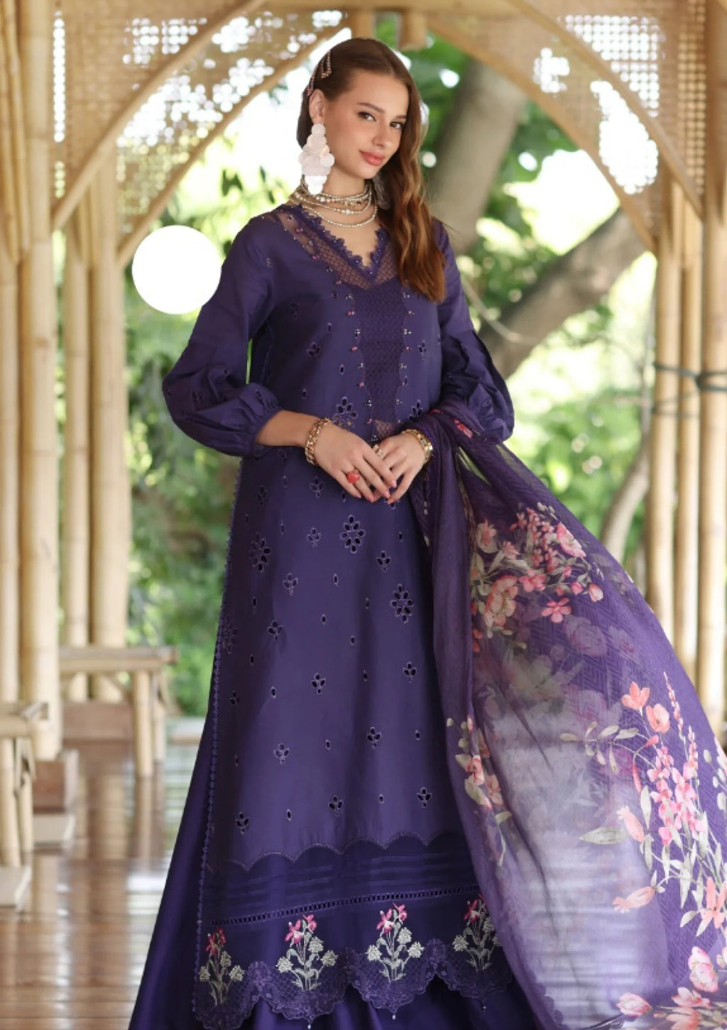 A woman models a deep purple traditional outfit with eyelet embroidery, featuring a long kurta, matching skirt, and a sheer floral dupatta, set against an arched wooden corridor with green foliage in the background.